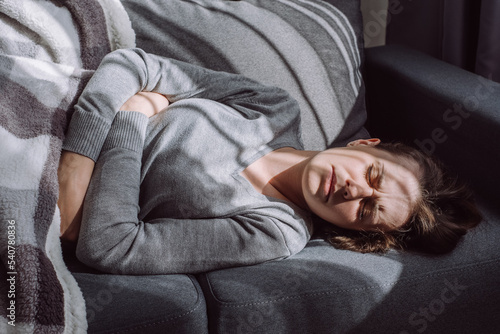 Unhealthy woman struggle with painful ache lying on couch at home. Sad unhappy young female suffering from menstrual pain, feeling sick to stomach, holding belly, having abdominal cramps during period