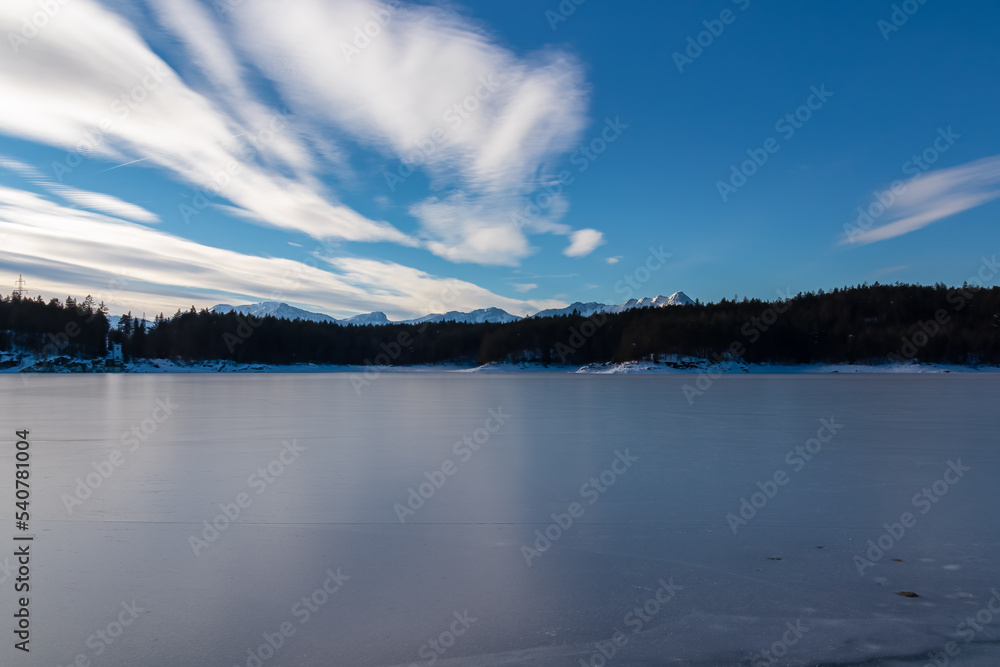 Fototapeta premium Scenic view of frozen alpine lake Forstsee, Techelsberg, Carinthia (Kaernten), Austria, Europe. Breathing fresh cold air. Winter wonderland. Snow capped Karawanks mountain range, Mittagskogel (Kepa)