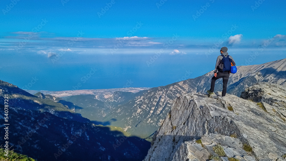 Rear view of man with climbing helmet on cloud covered mountain summit of Skala Mount Olympus, Mt Olympus National Park, Macedonia, Greece, Europe. View of rocky ridges and Mediterranean Aegean Sea