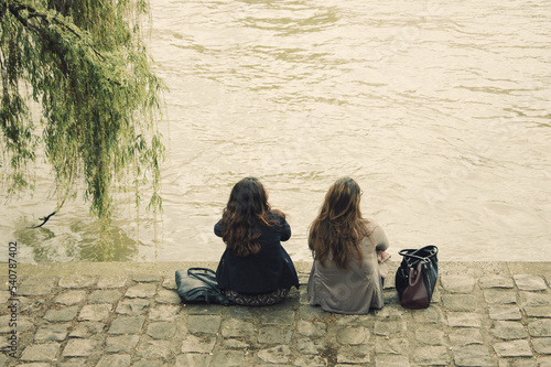Two young women relaxing at Seine river embankment in cloudy day. Paris, France Back view. Moody.