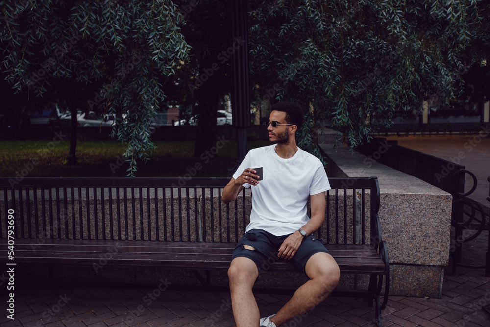 African american man in a white t-shirt sits on a bench and drinks coffee. Mock-up. African american man in a white t-shirt sits on a bench and drinks coffee. Mock-up.