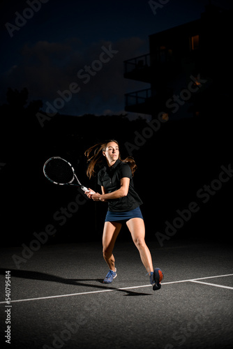 Great view on active woman with racket playing tennis on outdoor court at evening.