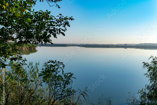 View of the river with banks overgrown with reeds and illuminated by the morning sun against a cloudless blue sky
