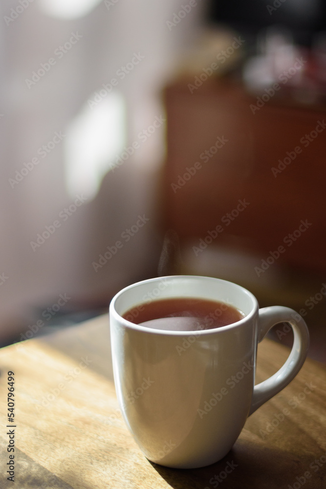 A mug full of coffee or tea set on a wooden table, morning sun and in the background light pink curtains A mug full of coffee or tea set on a wooden table, morning sun and in the background light pink curtains