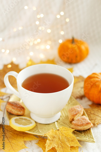 Creative mood. A cup of tea with lemon, cookies, autumn leaves and pumpkin on a white plaid with lights. Cozy autumn composition. Autumn mood concept.
