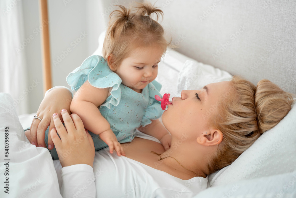 Mother and baby girl are lying in bed, enjoying time together. Mom keeps pacifier in her mouth. Family time concept Mother and baby girl are lying in bed, enjoying time together. Mom keeps pacifier in her mouth. Family time concept