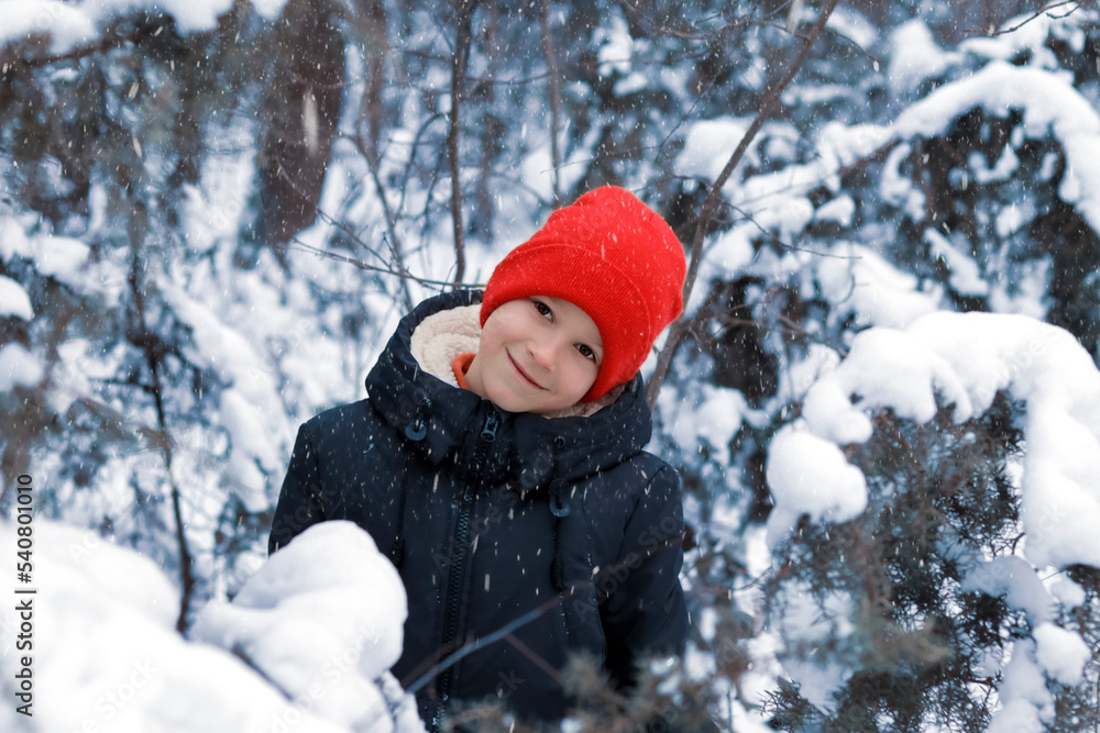 A happy smiling boy in a black down jacket and a red knitted hat plays and runs in a beautiful snowy winter park for Christmas A happy smiling boy in a black down jacket and a red knitted hat plays and runs in a beautiful snowy winter park for Christmas