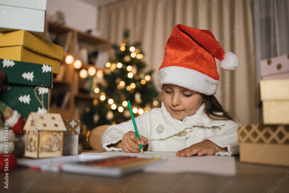 Letter for Santa. Cute little caucasian girl in red hat sitting at table and writing wish list of presents for Christmas in decorated room. Letter for Santa. Cute little caucasian girl in red hat sitting at table and writing wish list of presents for Christmas in decorated room.