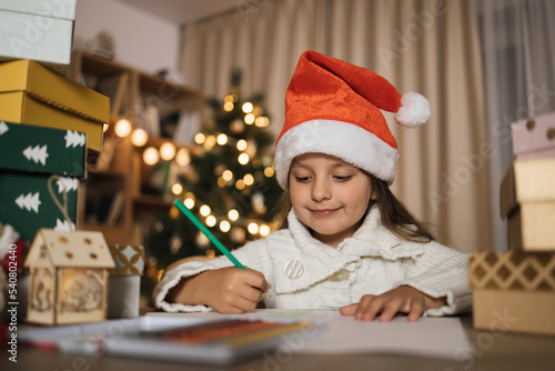 Letter for Santa. Cute little caucasian girl in red hat sitting at table and writing wish list of presents for Christmas in decorated room.