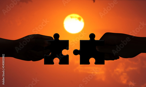 Two Hands Connecting 2 Puzzle Pieces Together in front of sunset background. Concept of Teamwork Support, Cooperative work and business association. 