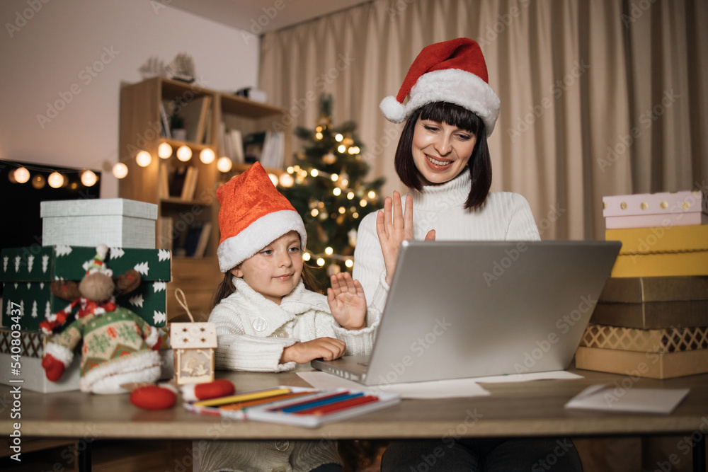 Young caucasian mother and her cute little daughter sitting at table and chatting with somebody using laptop in living room at home decorated with Christmas tree. Young caucasian mother and her cute little daughter sitting at table and chatting with somebody using laptop in living room at home decorated with Christmas tree.