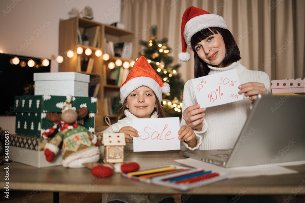 Concept of holiday discount. Young lovely mother and cute charming daughter kid in red hats holding letter with sale inscription at decorated with Christmas tree living room. Concept of holiday discount. Young lovely mother and cute charming daughter kid in red hats holding letter with sale inscription at decorated with Christmas tree living room.