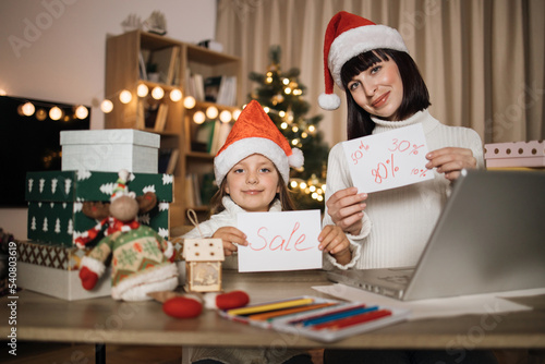 Concept of holiday discount. Young lovely mother and cute charming daughter kid in red hats holding letter with sale inscription at decorated with Christmas tree living room.