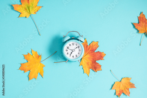 Alarm clock on blue background and autumn colorful leaves. Daylight saving time end. Fall leaves. Autumn background. Back to school concept. Flat lay.