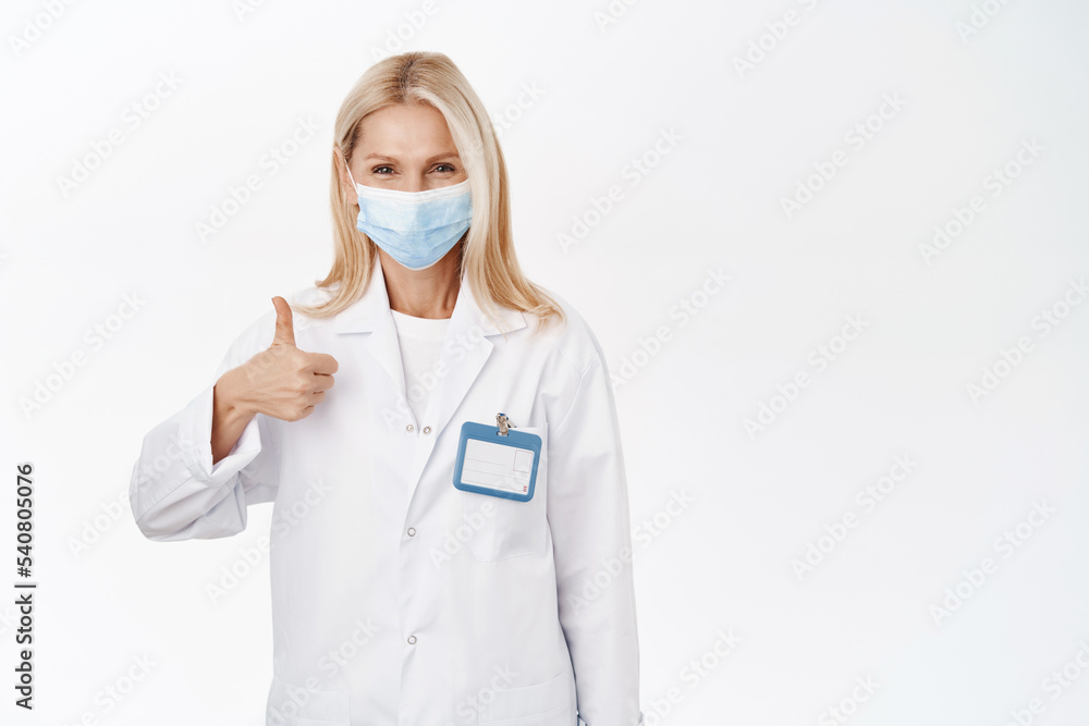 Smiling female doctor in medical face mask, shows thumbs up in approval, recommends smth, stands over white studio background Smiling female doctor in medical face mask, shows thumbs up in approval, recommends smth, stands over white studio background
