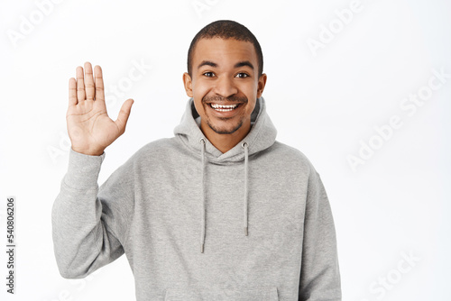 Portrait of friendly handsome man waves his hand, says hello, greets you with happy smile, stands over white background © Cookie Studio