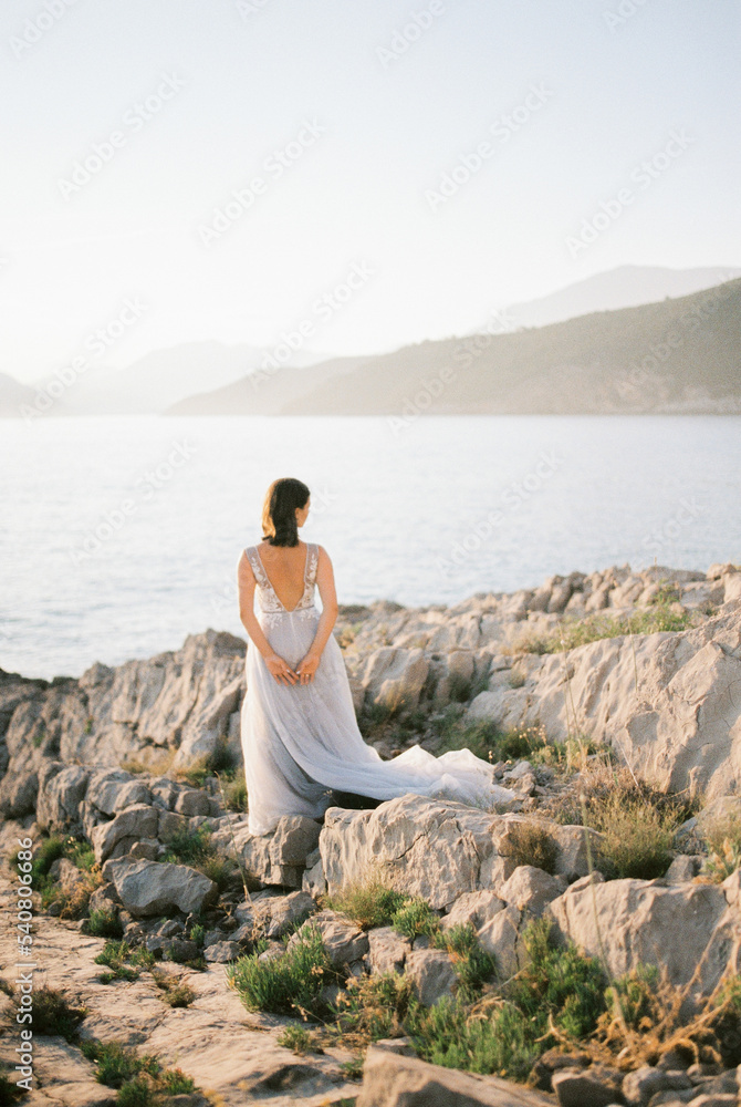 Bride in a white dress stands on a rocky seashore and looks at the mountains. Back view