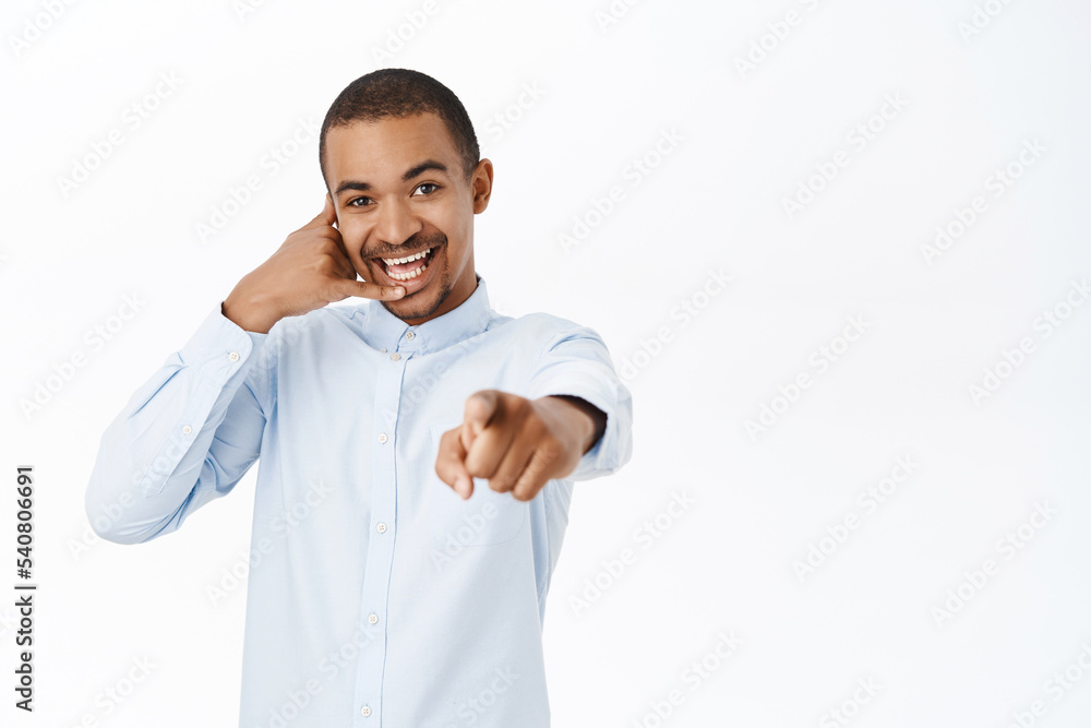 Smiling guy shows telephone gesture, points at camera, call us gesture, call center advertisement, standing over white background Smiling guy shows telephone gesture, points at camera, call us gesture, call center advertisement, standing over white background