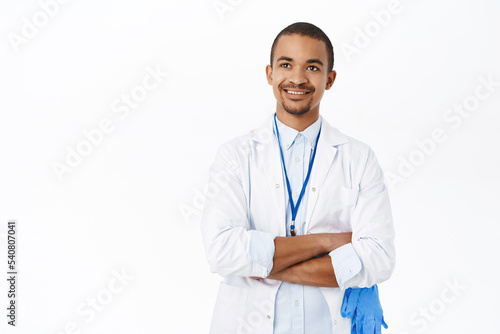 Smiling young doctor, intern in white coat, looking happy and confident, standing over white background