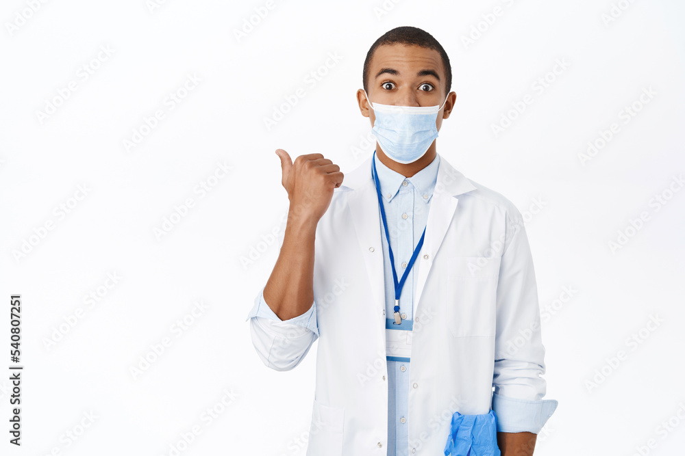 Surprised doctor in medical mask, points left, shows interesting advertisement, clinic promo offer, stands over white background Surprised doctor in medical mask, points left, shows interesting advertisement, clinic promo offer, stands over white background