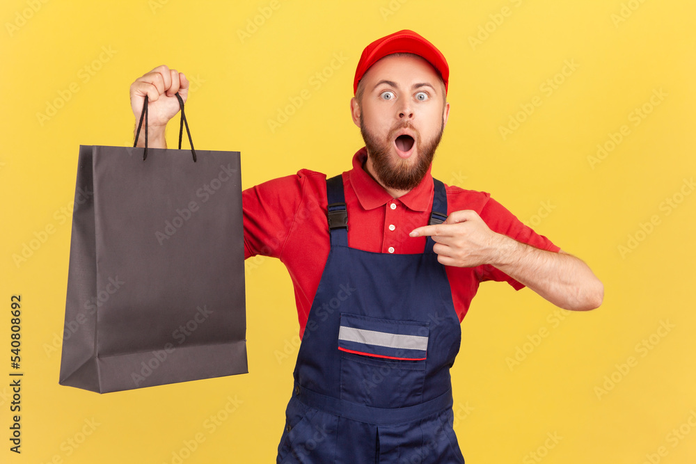 Amazed excited delivery man wearing blue uniform pointing at black shopping bag, delivering order to client, expressing astonishment, huge sale. Indoor studio shot isolated on yellow background