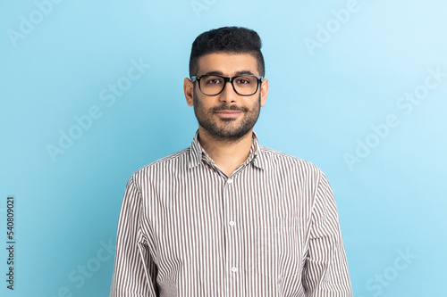 Portrait of smiling positive pleased businessman in optical eyeglasses standing looking at camera with smile, wearing striped shirt. Indoor studio shot isolated on blue background. © khosrork