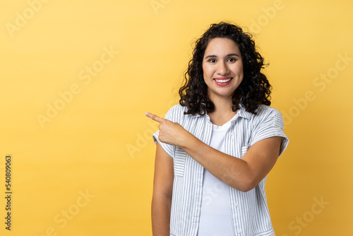 Portrait of smiling satisfied beautiful woman with dark wavy hair standing pointing aside with finger, showing copy space for advertisement. Indoor studio shot isolated on yellow background. photo