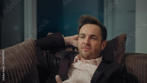 A young guy in a suit sits on a sofa at home and smiles.