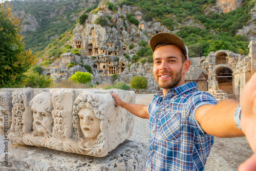 Swarthy caucasian european guy in plaid shirt smiles rejoices, takes selfie points with his hand show at the ancient unique landmarks Turkey, Antalya Demre, Myra, ancient lycian tomb casts faces. photo