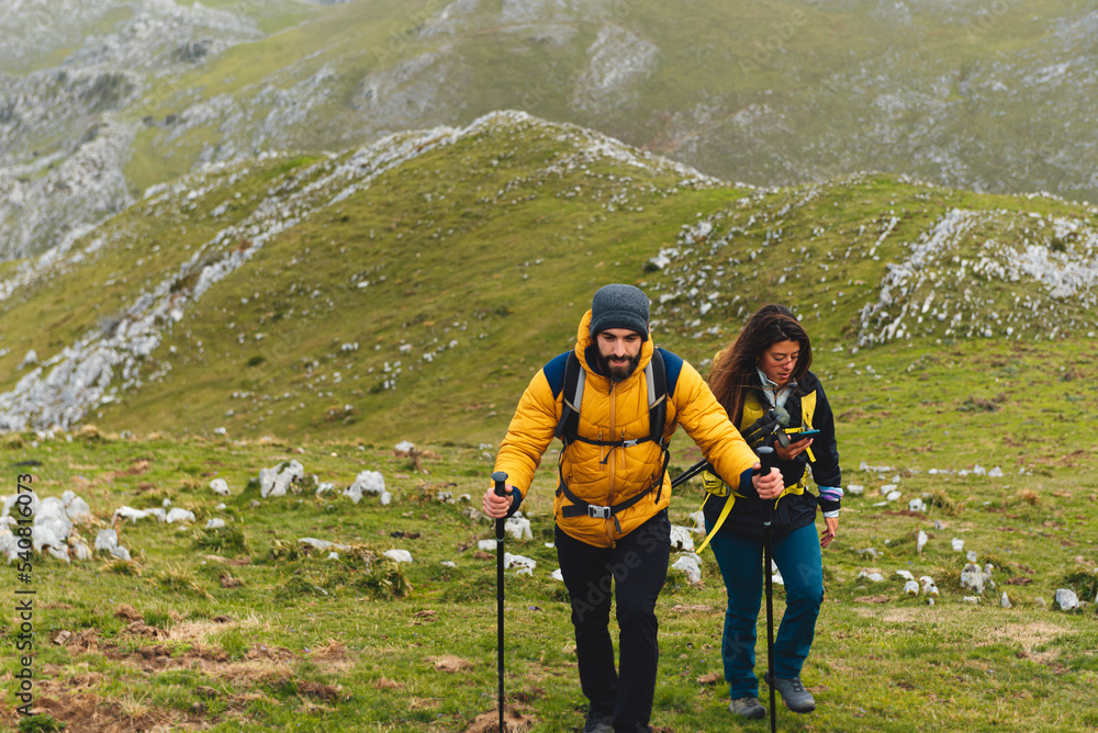 Couple of mountaineers, equipped with backpacks, trekking poles and using the mobile phone to watch the track, ascending a mountain. sports and outdoor activities.