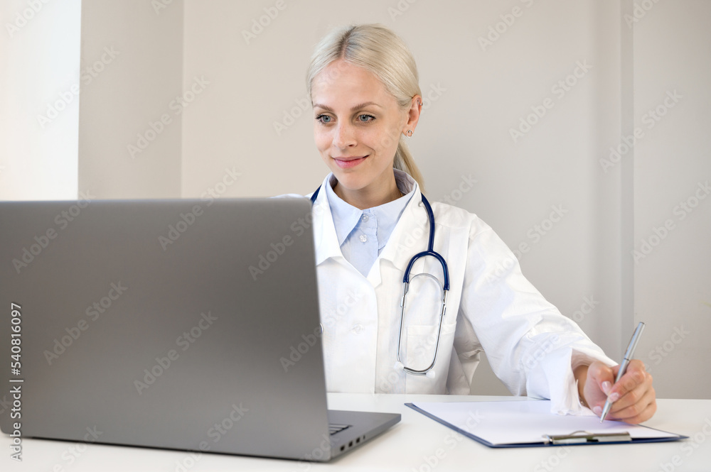 Portrait of confident young Caucasian female doctor in white medical uniform sit at desk filling out the forms looking at laptop screen Portrait of confident young Caucasian female doctor in white medical uniform sit at desk filling out the forms looking at laptop screen