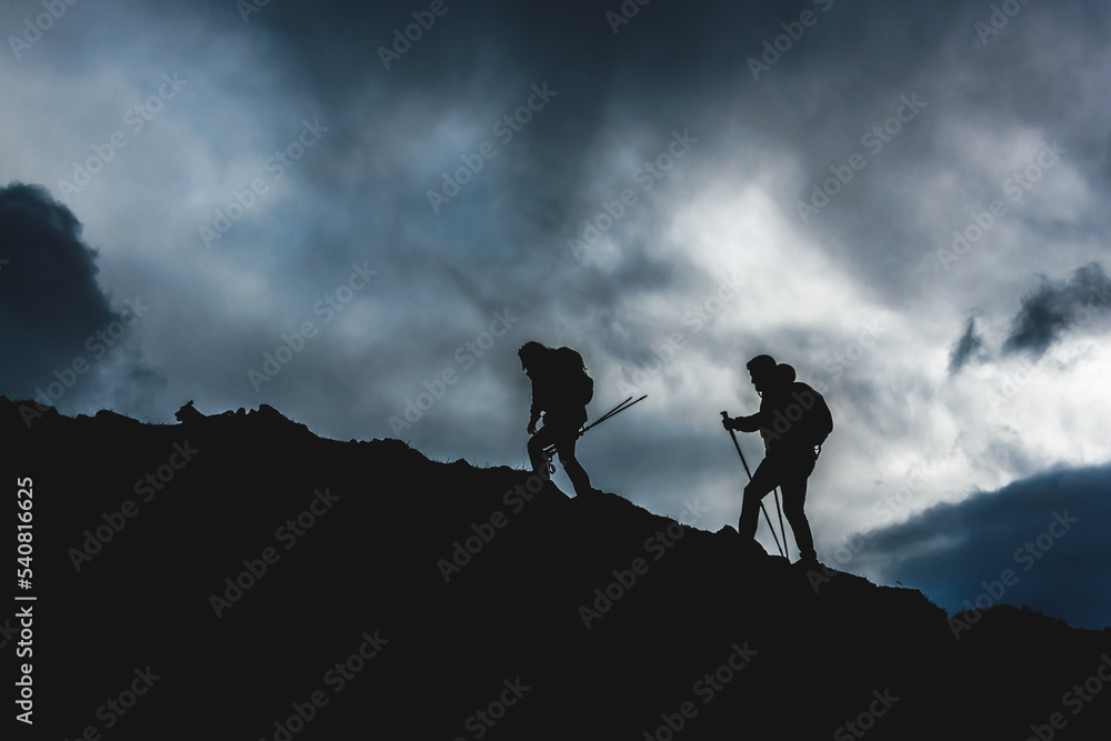 Underexposed photograph of two people in silhouette hiking up a mountain. couple ascending a mountain peak. sports and outdoor activities