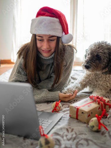 Christmas vacations online. Happy woman making a video call with her family or friends. Young woman using laptop computer lying on the floor with her dog on a blanket with gifts. Virtual meeting photo