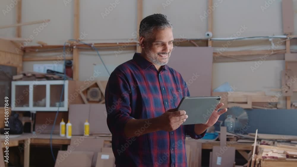 Mid adult man smiling and scrolling digital tablet while working in woodworking factory Mid adult man smiling and scrolling digital tablet while working in woodworking factory