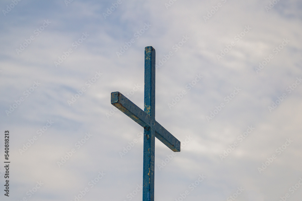 Uma cruz na cor azul com o céu nublado ao fundo.
