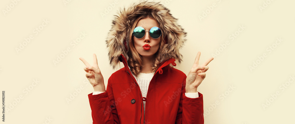 Portrait of beautiful young woman blowing her lips with red lipstick sending sweet air kiss wearing jacket with fur hood on blank background