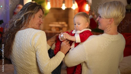 Back view two happy Caucasian women playing with pretty baby girl talking smiling. Relaxed cheerful mother and aunt enjoying Christmas eve with toddler indoors at home. New Year concept photo