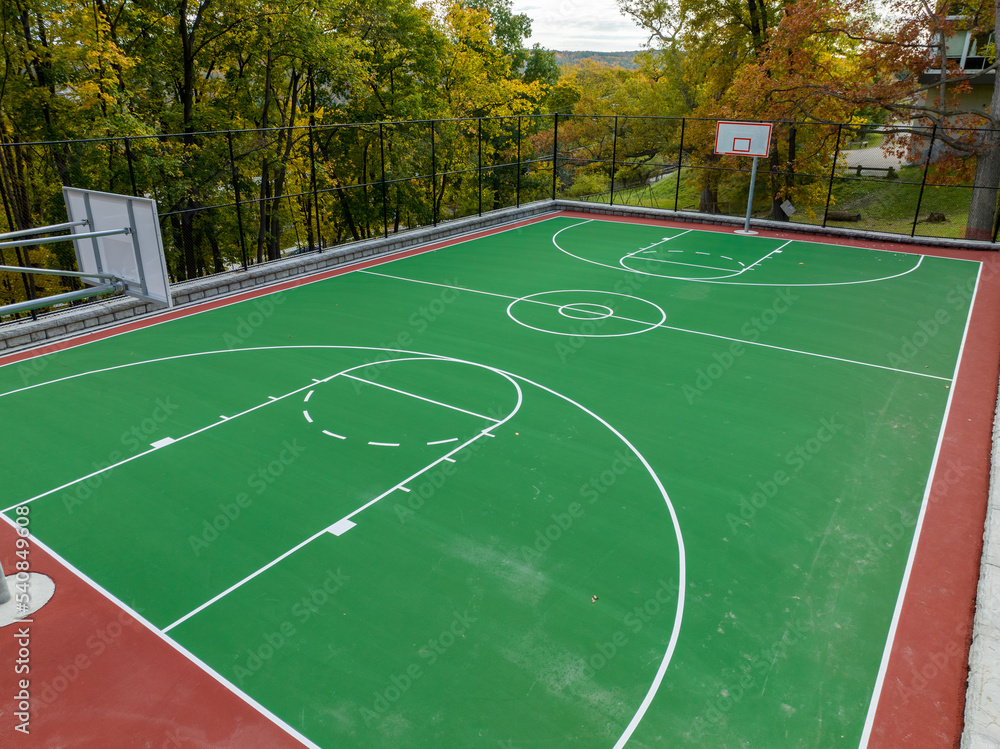 Aerial photo of a green and red outdoor basketball court at school playground.  Court includes retaining walls and black vinyl coated chain link fence.	