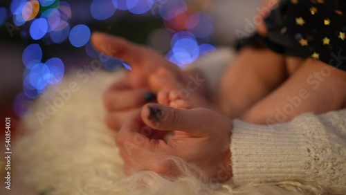 Close-up female hands tickling tiny baby feet on soft carpet at background of Christmas lights. Unrecognizable Caucasian loving mother playing with toddler daughter at home on New Year's eve