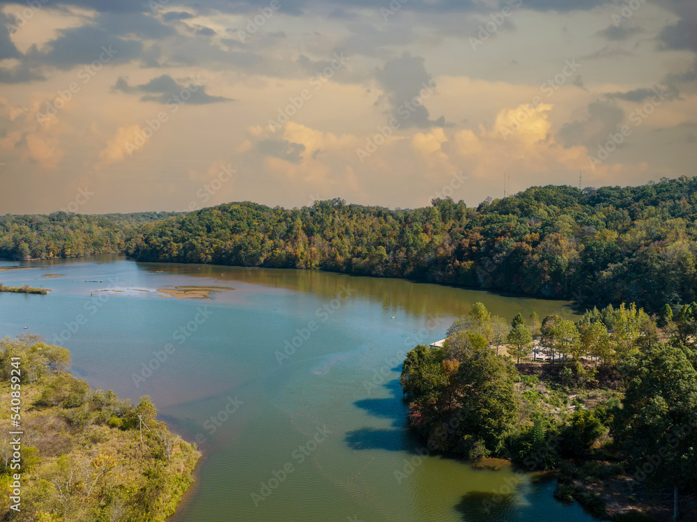 Naklejka premium Morgan Fall Overlook Park on sunny Fall day. Aerial river view with Fall and Autumn colored trees. Drone shot. Rolling storm clouds