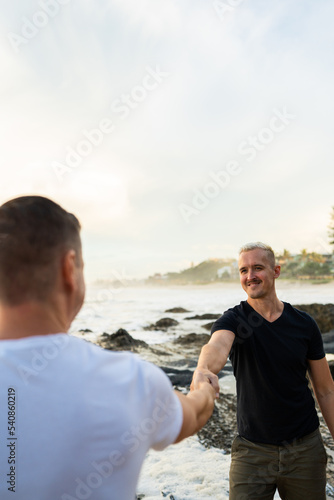 gay couple arm outstretched looking at each other with smile on beach