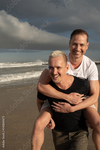 Gay couple piggy backing on beach smiling close up