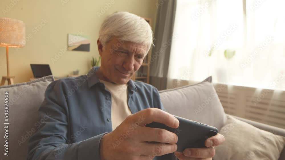 Handheld shot of excited old man playing game on smartphone sitting on sofa at home Handheld shot of excited old man playing game on smartphone sitting on sofa at home