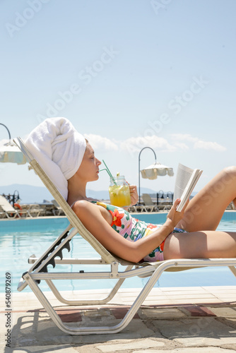 Portrait of beautiful young woman enyojing hot day, having cocktail and reading a book on the swimingpool. Relaxing summer vacation concept. photo