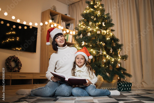 Merry Christmas and Happy Holidays. Young happy mother and little cute daughter in santa hat reading magical fairy tale book sitting near decorating tree in living room. Loving family indoors.