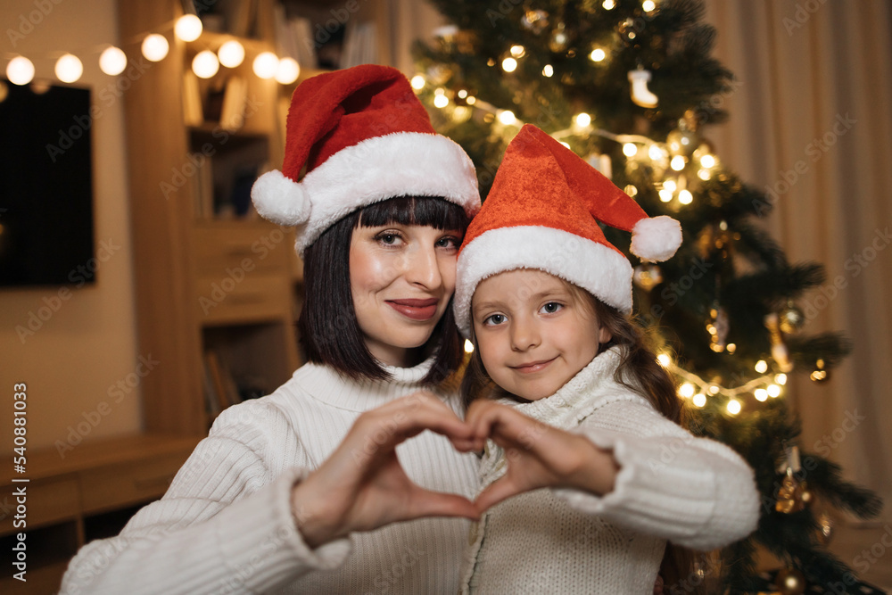 Close up of lovely family of two, beautiful young mother and her cute little daughter in warm knitted white sweaters making heart-shaped sign with their hands in front of decorated Christmas tree. Close up of lovely family of two, beautiful young mother and her cute little daughter in warm knitted white sweaters making heart-shaped sign with their hands in front of decorated Christmas tree.