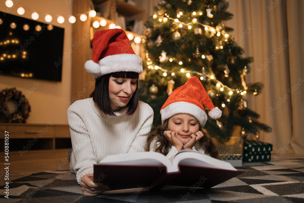 Pretty young mother in santa hat reading magical fairy tale book to her daughter lying on floor near Christmas tree, merry Christmas. Pretty young mother in santa hat reading magical fairy tale book to her daughter lying on floor near Christmas tree, merry Christmas.