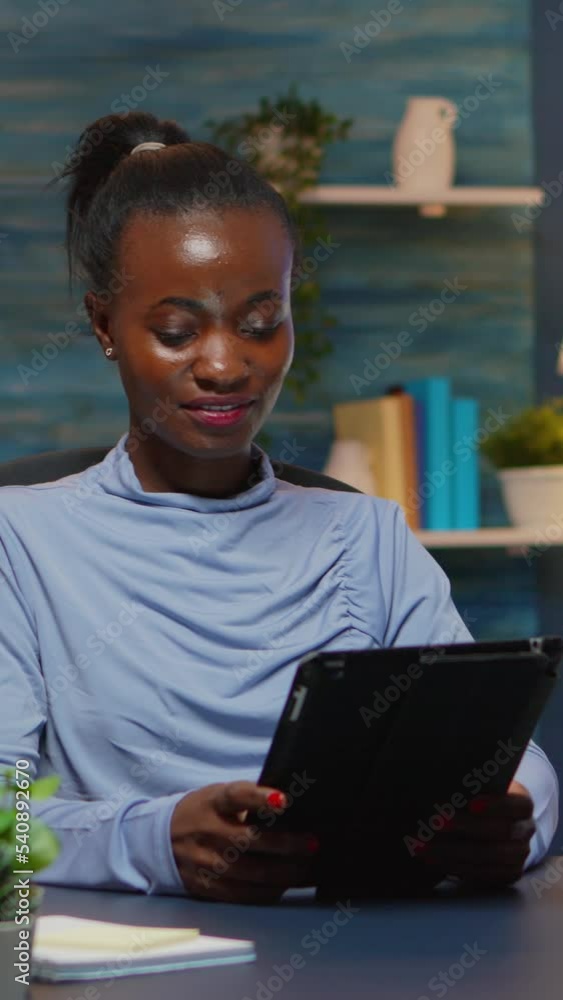 Vertical video: African american businesswoman taking break using tablet late at night sitting in modern living room. Busy focused employee using modern technology network wireless doing overtime