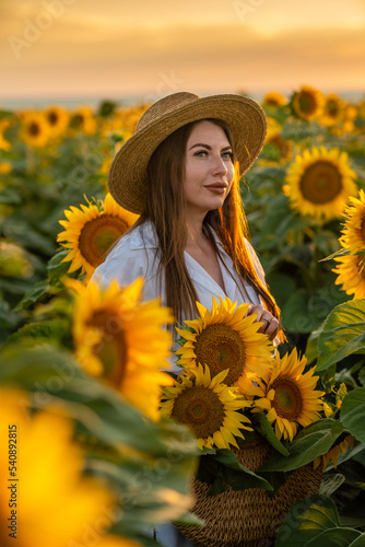 A girl in a hat on a beautiful field of sunflowers against the sky in the evening light of a summer sunset. Sunbeams through the flower field. Natural background.