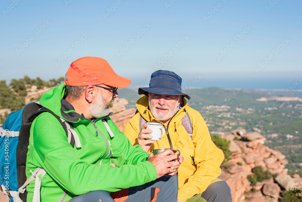 Two happy elderly trekkers sitting on the summit with a cup